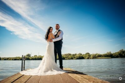 Bride and groom on jetty by river under blue sky.