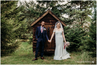 Couple outside wooden cabin, wedding attire, forest background.