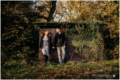 Couple standing by rustic shed in autumn sunlight.