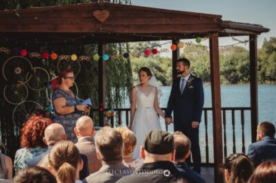 Outdoor lakeside wedding ceremony with couple and guests.