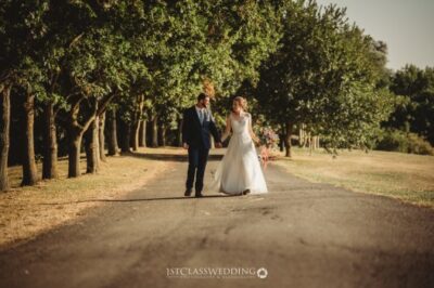 Couple walking hand-in-hand on tree-lined path at wedding.