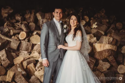 Bride and groom smiling in front of woodpile.