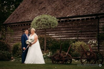 Wedding couple posing by rustic barn and vintage cart.