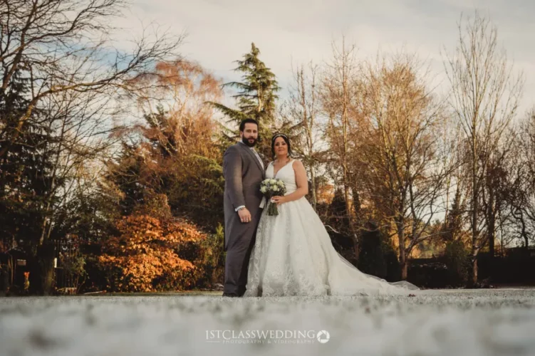 Couple in wedding attire outdoors with autumn trees.