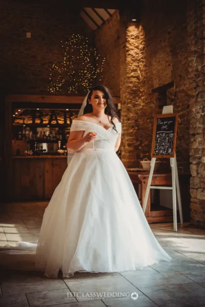 Bride in white dress holding champagne in rustic setting.