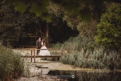 Bride and groom by serene lake with woodland backdrop.