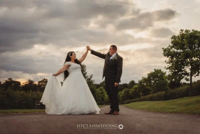 Bride and groom dancing on path under cloudy sky.