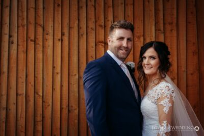 Couple posing in wedding attire, wooden backdrop.