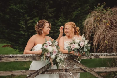 Two brides laughing with bouquets by wooden fence.