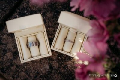 Wedding rings in open boxes with pink flowers nearby.