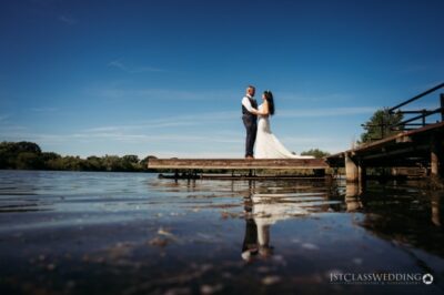 Bride and groom embracing on lakeside dock, clear sky.
