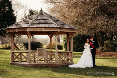 Bride and groom posing by wooden gazebo outdoors.