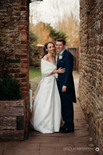 Bride and groom embracing beside old brick wall.