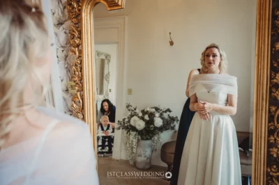 Elegant bride in mirror with baby and woman behind.