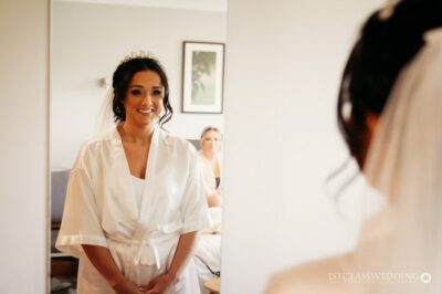 Bride smiling, preparing in robe, reflection in mirror.