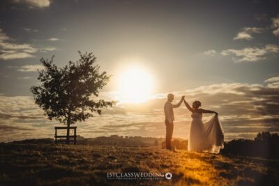 Couple holding hands at sunset in countryside wedding scene.