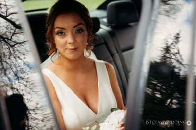 Bride in car holding bouquet, reflective photography.