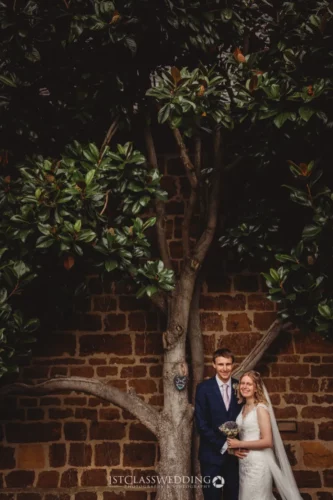 Couple at wedding with tree and brick wall backdrop.
