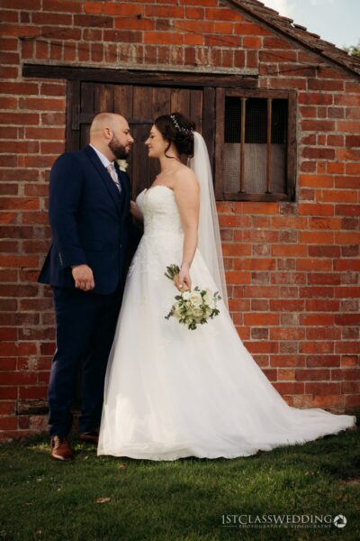 Couple posing in wedding attire by a brick building.