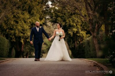 Bride and groom holding hands on tree-lined path.