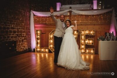 Bride and groom celebrating with illuminated 'LOVE' sign.