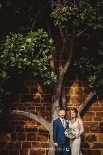 Wedding couple posing by brick wall and tree.