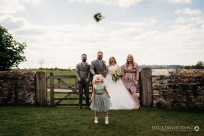 Bride tossing bouquet, wedding guests in countryside setting.
