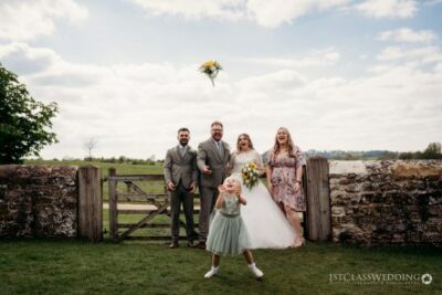 Bride tossing bouquet, joyful wedding guests in countryside.