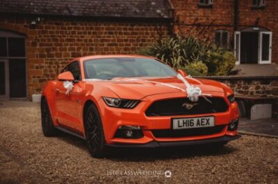 Orange Ford Mustang wedding car with white ribbons.