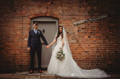 Couple in wedding attire holding hands before brick facade.