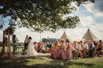 Outdoor wedding ceremony under tree with guests and tipis.