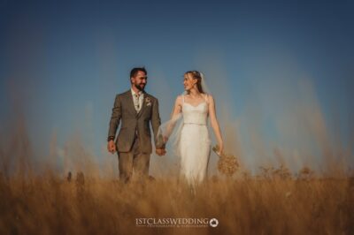 Bride and groom walking in field at sunset.