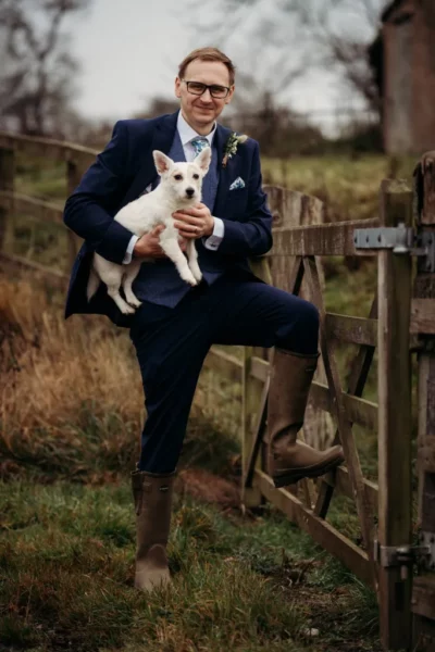Man in suit holding dog by wooden gate.