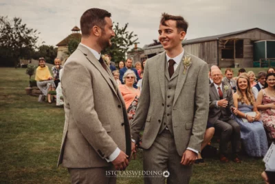 Two men in suits smiling at rustic wedding ceremony.