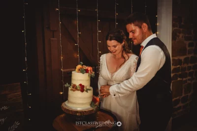 Couple cutting wedding cake with fairy lights behind.