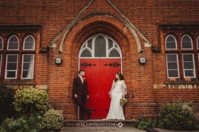 Couple by red door at brick wedding venue.