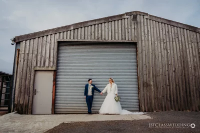 Bride and groom holding hands by industrial building.