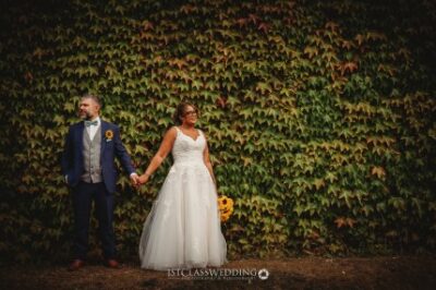 Bride and groom holding hands before ivy-covered wall.