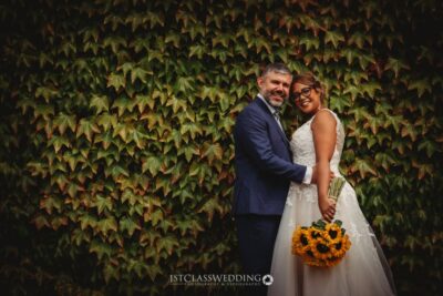 Bride and groom embracing, ivy backdrop, sunflower bouquet.