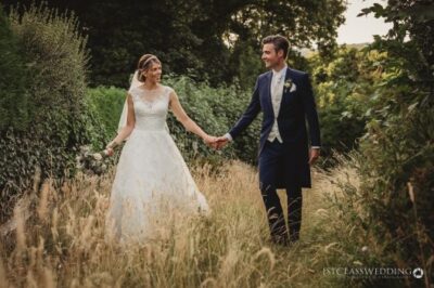 Bride and groom walking hand in hand in meadow.