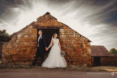 Bride and groom holding hands by rustic stone barn.