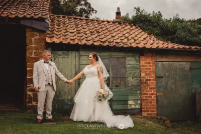 Bride and groom holding hands by rustic building.