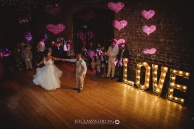 Couple dancing at wedding with illuminated 'LOVE' sign.