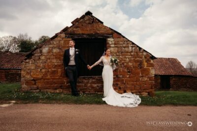 Newlyweds posing by rustic stone barn, countryside wedding.