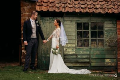 Bride and groom holding hands by rustic building.