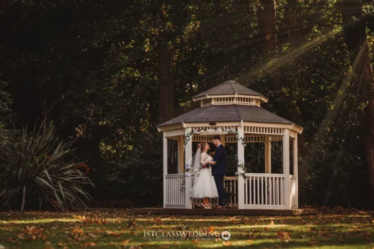 Wedding couple embracing in sunlit gazebo surrounded by trees.