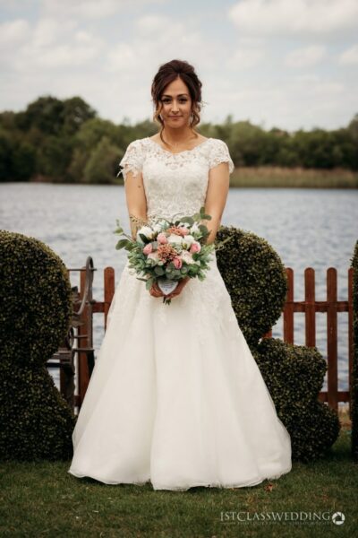 Bride in white dress holding bouquet by the lake.