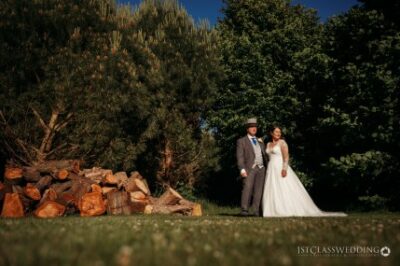 Couple in wedding attire outdoors with trees and firewood.