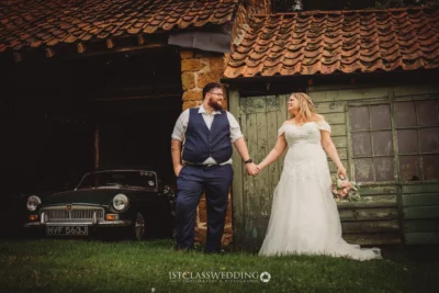 Wedding couple holding hands by vintage car and barn