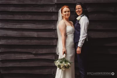 Two brides smiling, wedding attire, wooden backdrop.